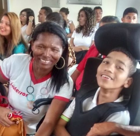 Mulher sorrindo sentada ao lado de rapaz jovem e sorridente sentado numa cadeira de rodas.