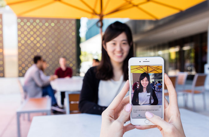 Um celular rodando o aplicativo Seeing AI apontando em direção a uma pessoa em um restaurante. O aplicativo sugere que ela é uma mulher de 25 anos com cabelo preto e parecendo alegre.
