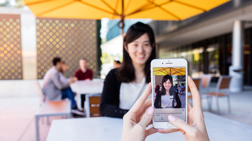 Um celular rodando o aplicativo Seeing AI apontando em direção a uma pessoa em um restaurante. O aplicativo sugere que ela é uma mulher de 25 anos com cabelo preto e parecendo alegre.